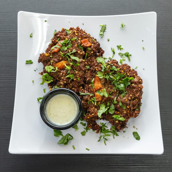 Two crispy brown fritters topped with cilantro and orange pieces, served with a side of white sauce.